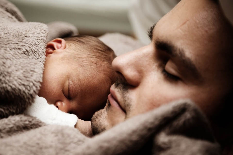 A baby curled up on their father's chest, with the father having his eyes closed, surrounded with a fluffy grey blanket.